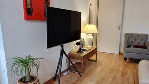 Een woonkamer met een flatscreen televisie op een standaard, een klein tafeltje met een lamp, een kamerplant en een bank.