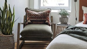A room with a wooden chair, a large window, a bedside table with a lamp, and a potted plant.