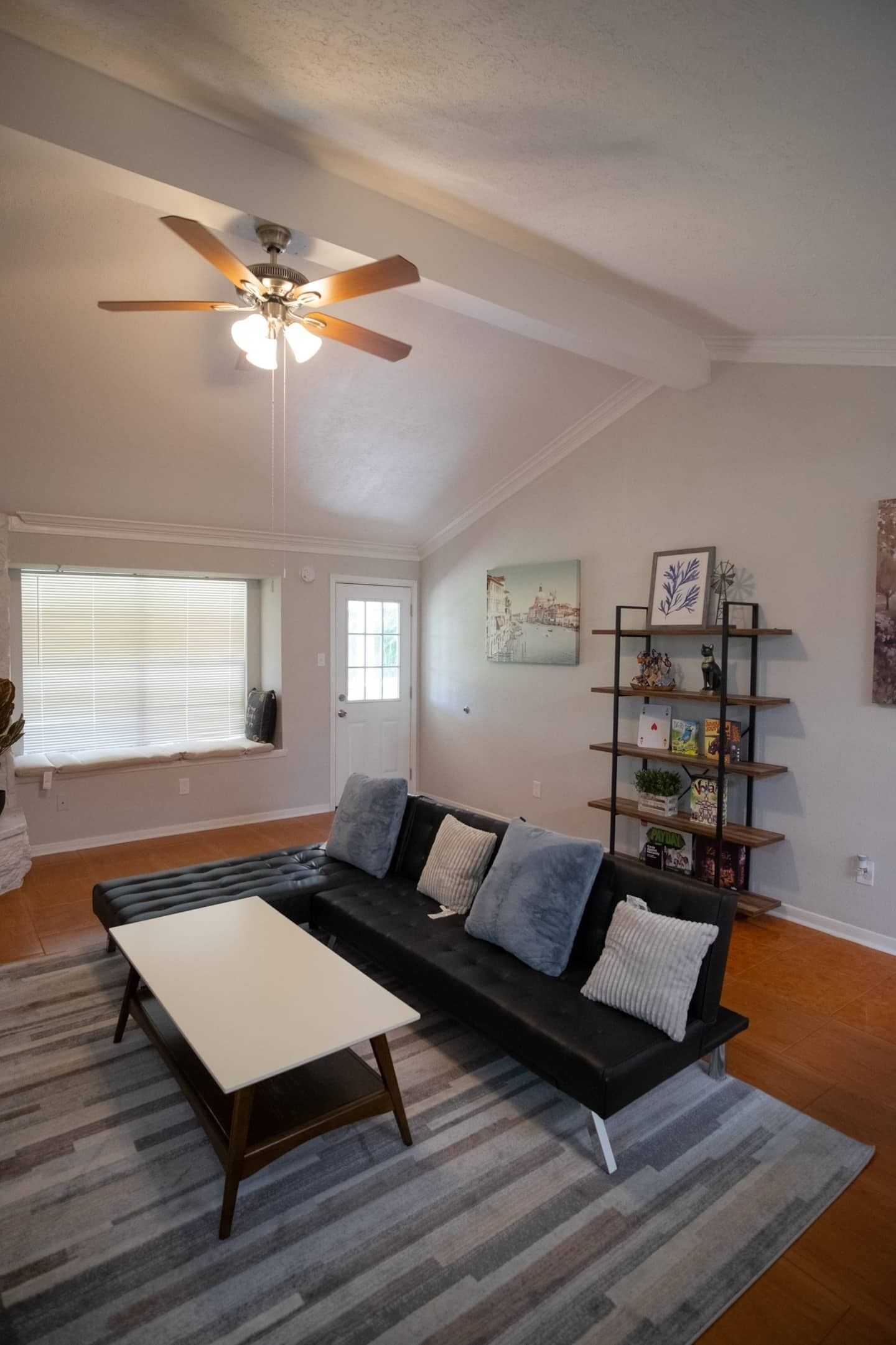 A living room with a black sectional sofa, a white coffee table, a ceiling fan, a bookshelf, and a window with blinds.