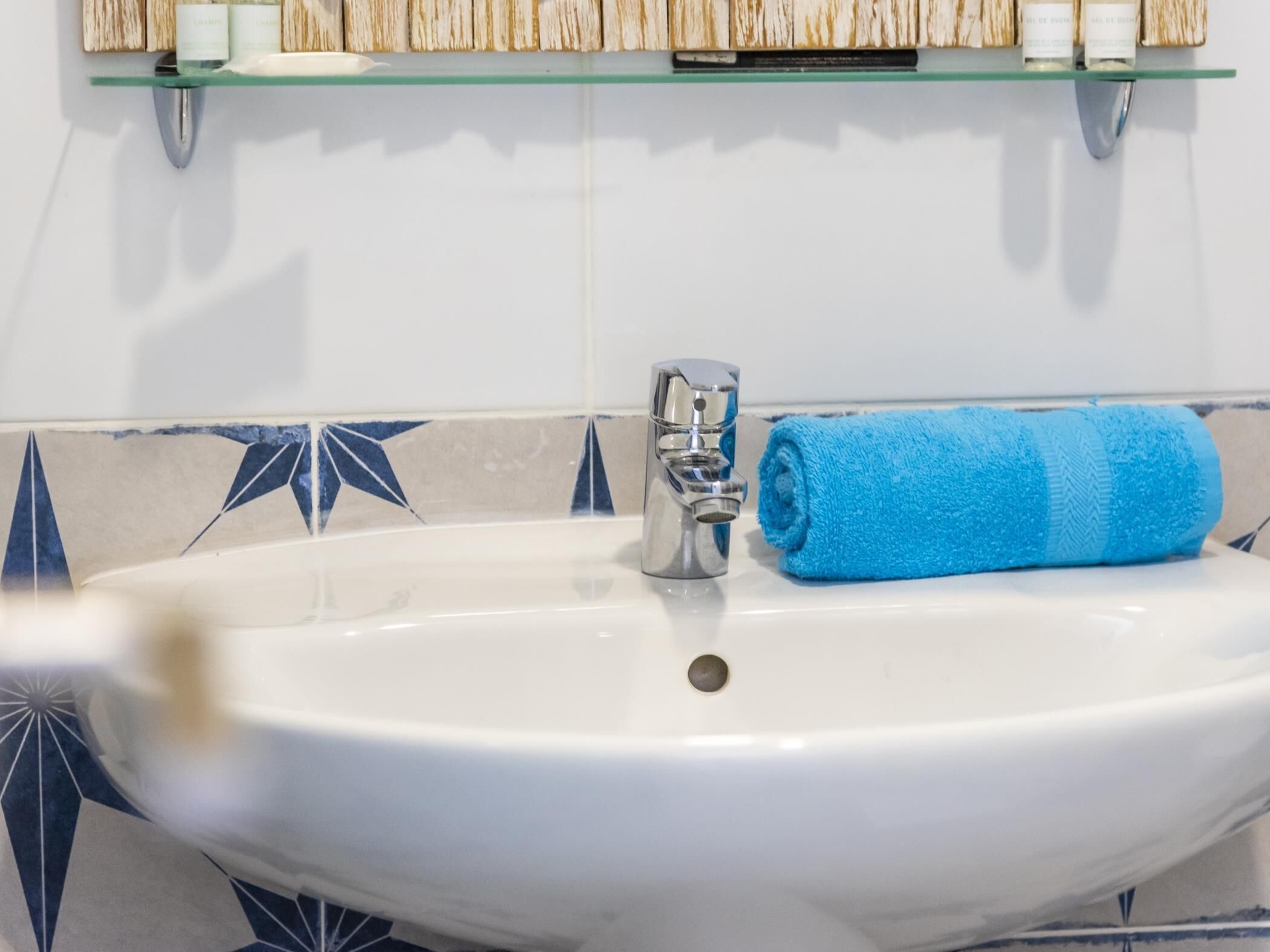 Un lavabo de bany amb una tovallola blava, una prestatgeria de vidre a sobre amb productes de tocador, i un frontal de rajoles amb un disseny.