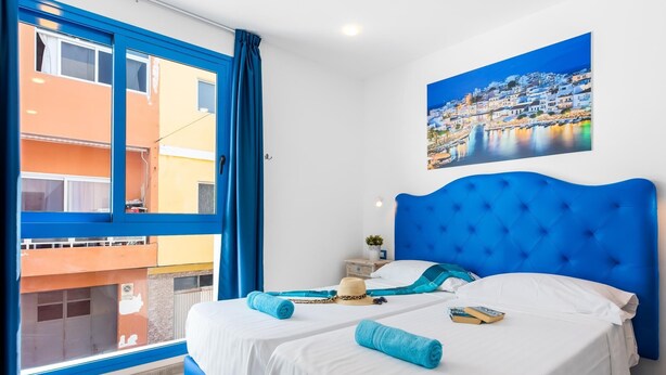 A hotel room with two beds, a large window with blue curtains, a painting of a coastal town, and a view of a building outside.