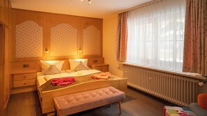 Een slaapkamer met een houten bed, nachtkastjes, een bankje, een radiator en een raam met gordijnen.