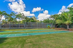 A tennis court with a green surface, surrounded by a chain-link fence, palm trees, and a clear blue sky.