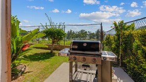 Ein Grill im Garten mit einem Maschendrahtzaun, Blumentöpfen und blauem Himmel.