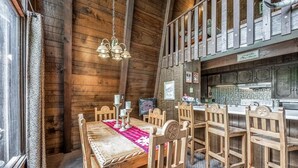 A wooden interior with a dining table set for four, surrounded by wooden chairs. A chandelier hangs from the ceiling, and a kitchen area is visible in the background. - Lazy Dog Lodge - hot tub, dog friendly, wifi (Ruidoso)