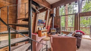 A rustic cabin interior with a wooden staircase, a fireplace, a sofa, and a large window overlooking trees. - Lazy Dog Lodge - hot tub, dog friendly, wifi (Ruidoso)
