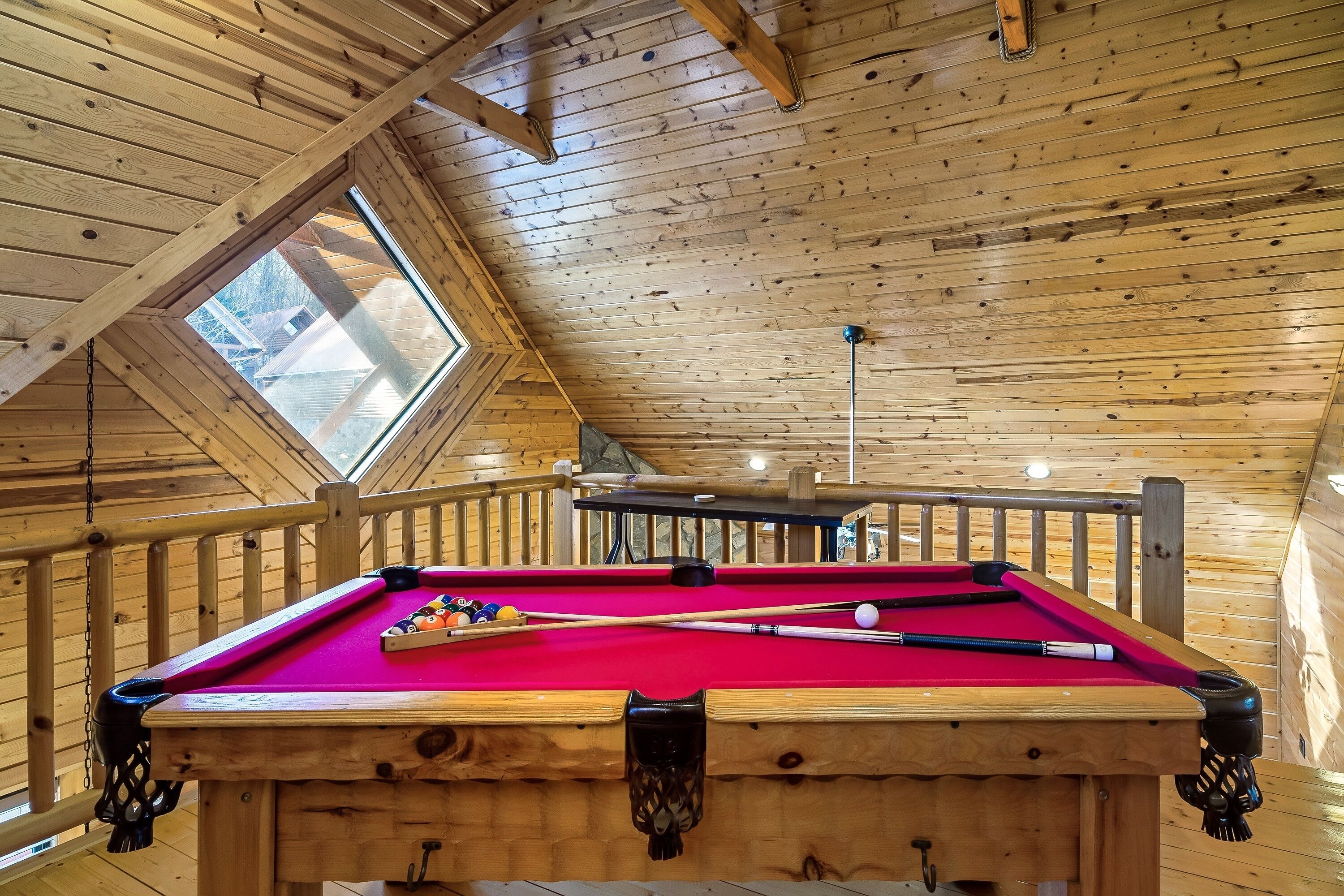 A wooden pool table with a red felt surface, surrounded by wooden railings and a sloped ceiling with skylights.