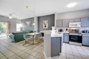 Una cucina moderna con mobili grigi, un'isola bianca ed elettrodomestici in acciaio inossidabile. C'è una zona pranzo con tavolo e sedie, e un divano verde sullo sfondo.