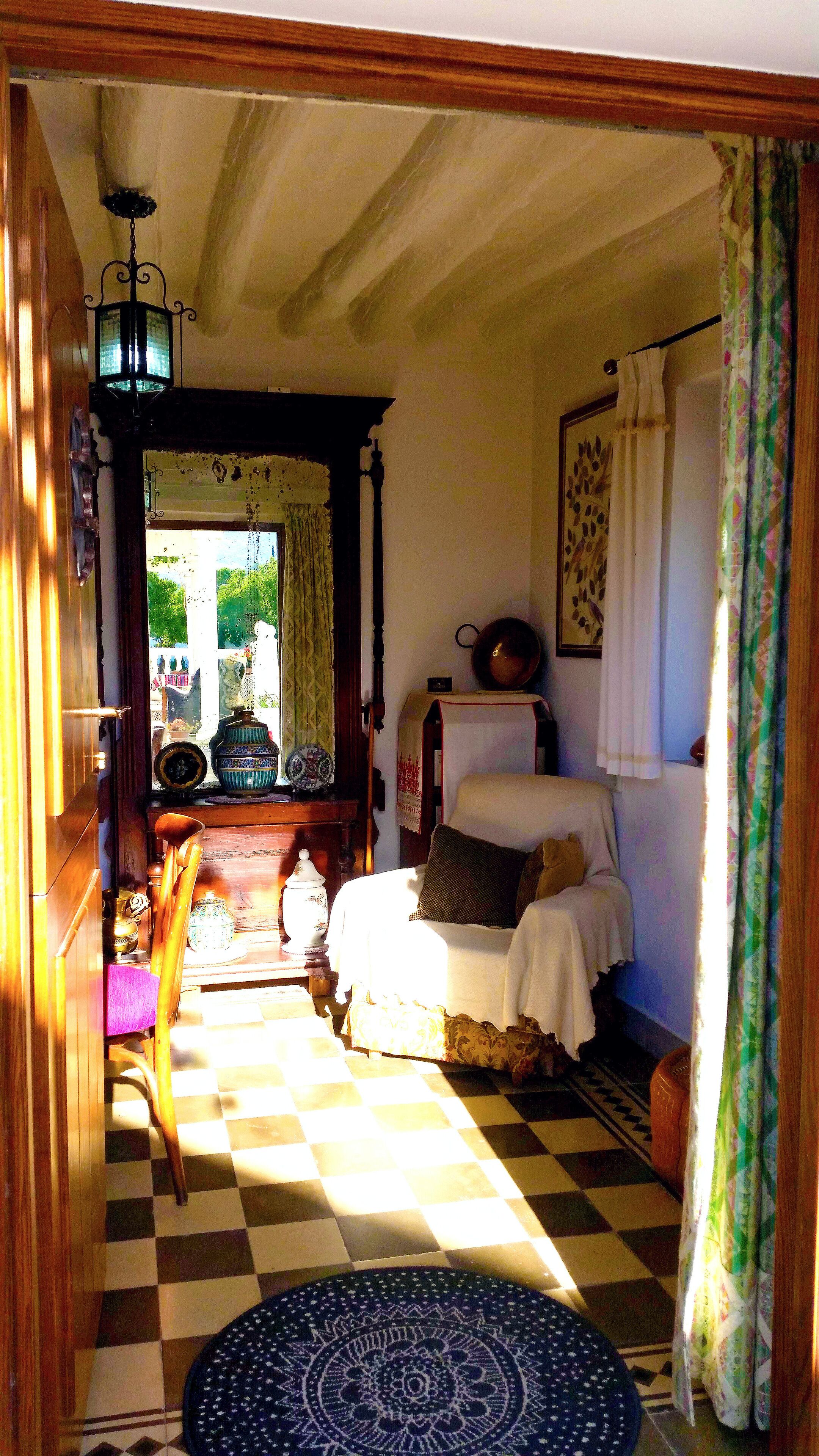 Un cuarto acogedor con piso a cuadros, una cama con cubrecama blanco, una silla, una mesa con un florero y una ventana con cortinas.