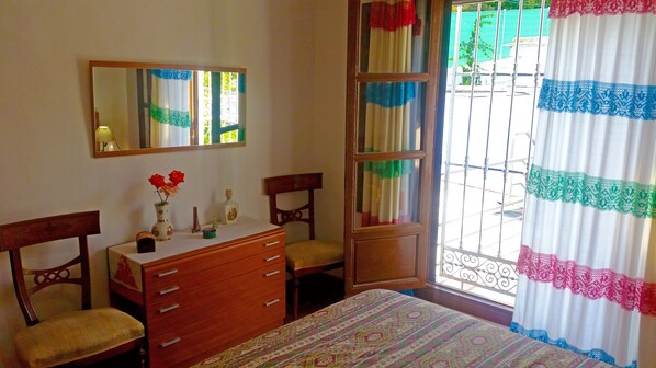 A bedroom with a bed, a dresser, two chairs, a mirror, and a window with curtains.