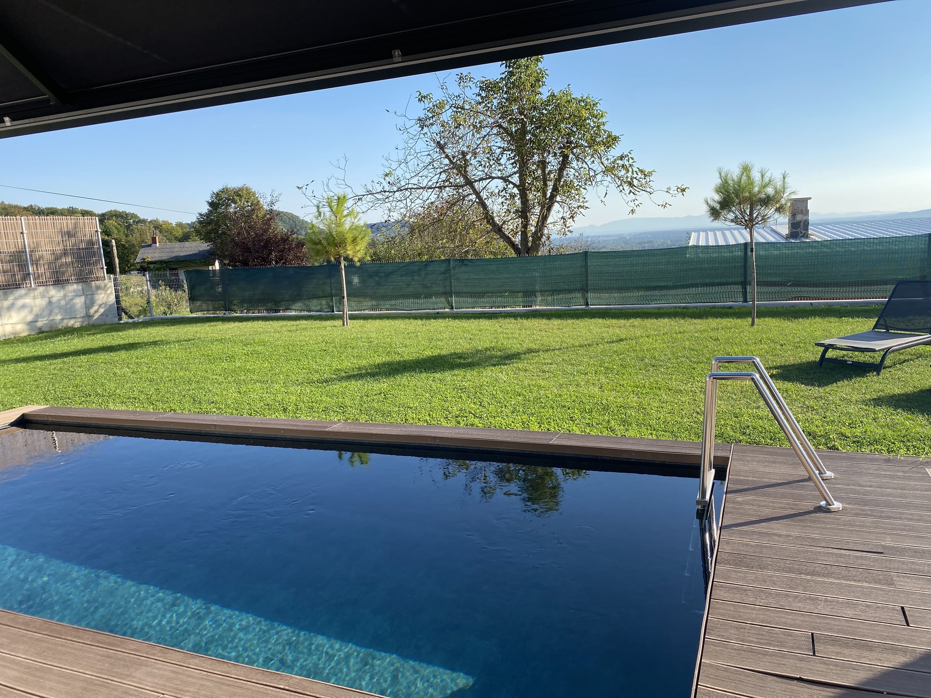 Una piscina con muelle de madera y vista a un jardín césped con un árbol y una cerca.