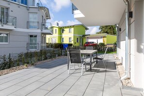 A patio area with a table and chairs, a red car in the background, and a building with balconies. - Domizil Meerzeit, App. 2 (Heringsdorf)