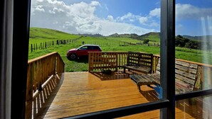 Une terrasse en bois avec un banc et un SUV rouge garé dans un champ herbeux, avec une clôture et des collines en arrière-plan.