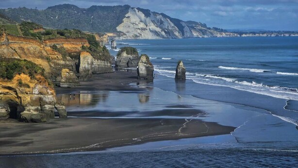 Un paysage côtier caractérisé par des formations rocheuses, une plage de sable et une chaîne de montagnes à l’horizon.