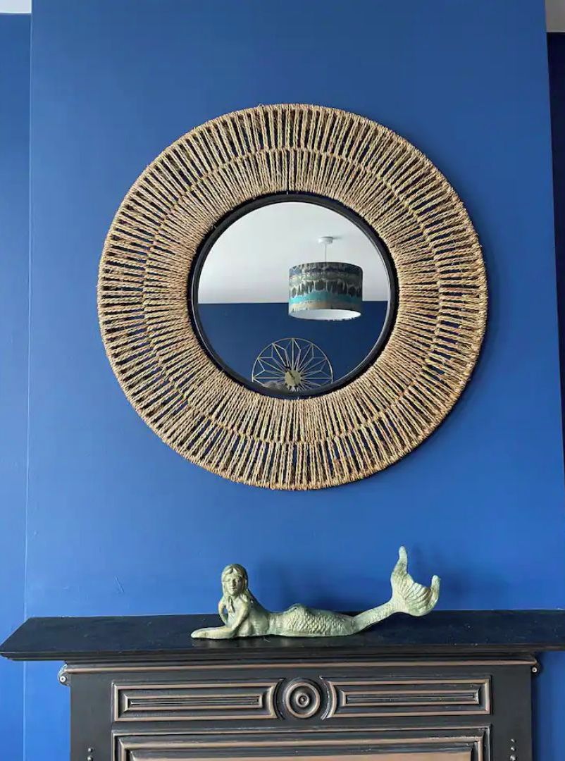 A circular mirror with a textured frame on a blue wall, a decorative mermaid figure on a black mantelpiece below, and a view of a building through the mirror.