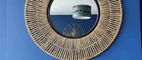 A circular mirror with a textured frame on a blue wall, a decorative mermaid figure on a black mantelpiece below, and a view of a building through the mirror.