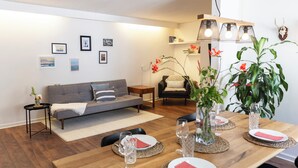 Un salon moderne avec un canapé, une table à manger pour deux personnes et une table basse. On peut y voir des tableaux encadrés au mur, une lampe et une plante.