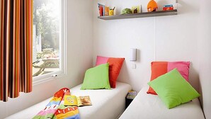 A room with two beds, colorful pillows, a window with curtains, and a shelf with various items. - Woodland Chalet in the Ardennes (Olloy-sur-Viroin)