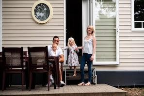 A family of four seated on a porch with a wooden chair and a bench, a window with a round mirror, and a door leading to the interior.