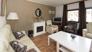 Een woonkamer met een witte bank, een witte salontafel, een open haard, een televisie en een raam met gordijnen.