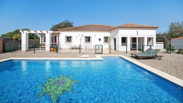 Une piscine avec un élément décoratif aquatique à l’eau d’un bleu limpide, un bâtiment blanc doté d’un toit en tuiles et un patio couvert.