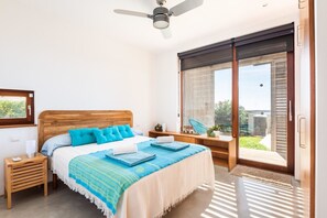 Un dormitorio con cabecera de madera, una cama con ropa de cama azul, un ventilador de techo, una ventana con vistas y una mesita de noche con lámpara.
