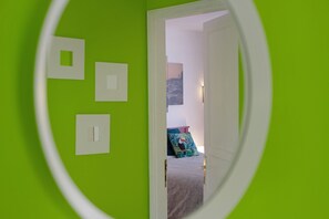 Une pièce avec un mur vert, une porte donnant sur une autre pièce et un lit agrémenté d’un coussin à motifs.