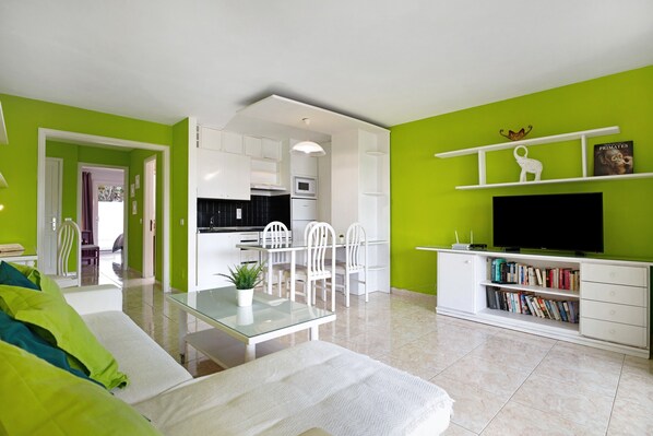 Un salon moderne avec une table basse en verre, un canapé, un coin repas avec des chaises, une cuisine avec des meubles blancs et une bibliothèque remplie de livres et d’un téléviseur.