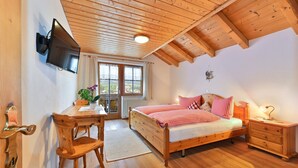 Ein Schlafzimmer mit Holzboden, einem Bett mit karierter Bettwäsche, einem Nachttisch mit Lampe, einem Holztisch mit Stuhl und einem an der Wand befestigten Fernseher.