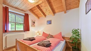 Una cama de madera con ropa de cama a cuadros, una ventana con cortinas, un radiador, una lámpara fijada a la pared y una planta en maceta.