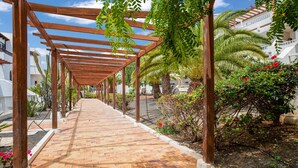 Un paseo techado con vigas de madera y un sendero empedrado, rodeado de exuberante follaje y plantas florecientes.