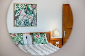 Une chambre à coucher avec un lit, des oreillers, une table de chevet, une lampe, une porte en bois et un tableau sur le thème des pays tropicaux.