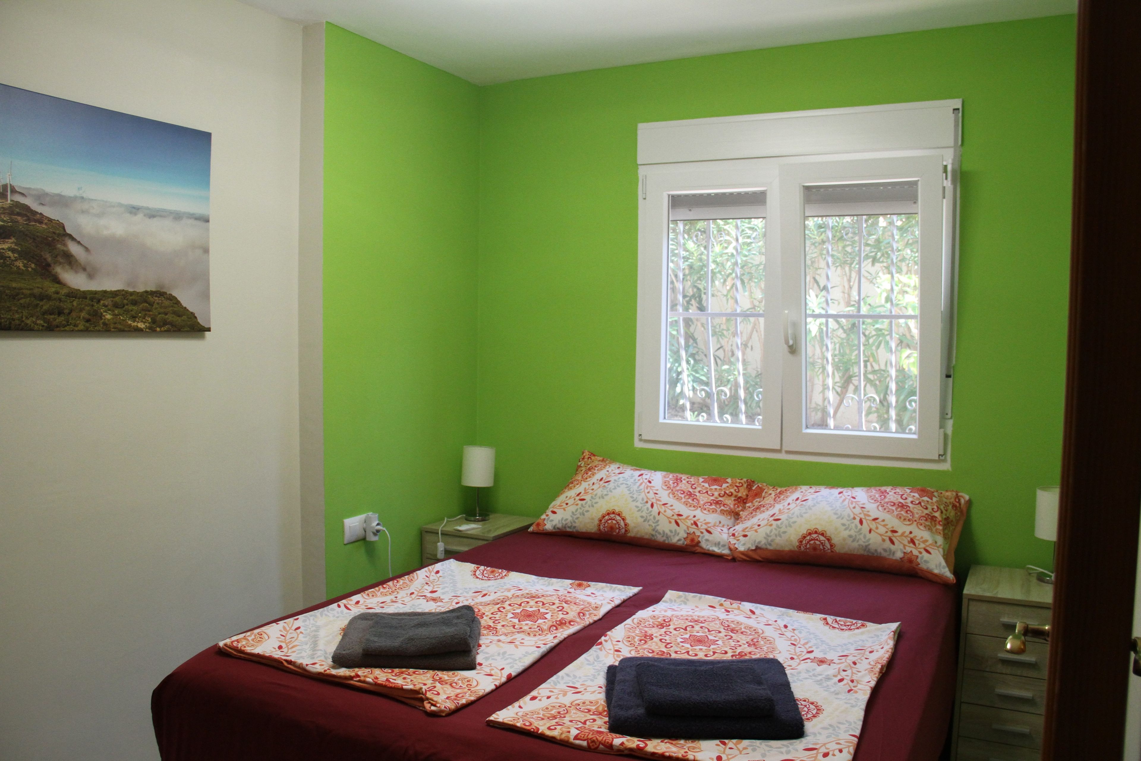 Una cama individual con colcha roja y dos almohadas, una mesita de noche con lámpara, y una ventana con vistas a la vegetación.