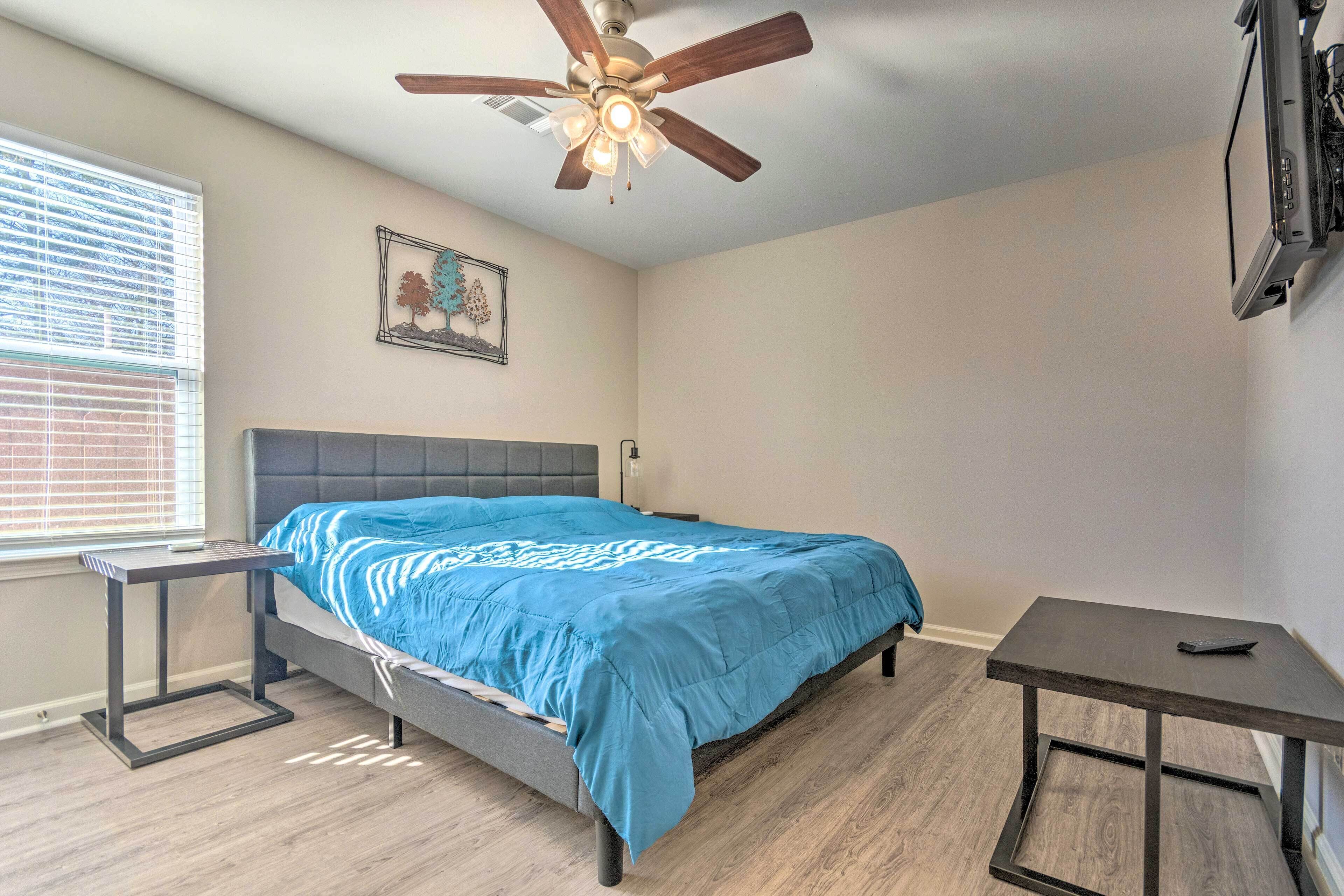 Une chambre à coucher avec un lit, un ventilateur de plafond, une fenêtre avec des stores et un téléviseur fixé au mur.