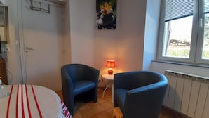 Une petite pièce avec deux fauteuils bleus, une petite table sur laquelle se trouve une lampe rouge, un tableau accroché au mur et une fenêtre avec des stores.