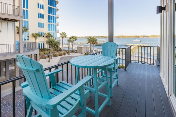 A balcony with turquoise Adirondack chairs and a small table, overlooking a body of water with a dock and a gazebo.
