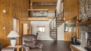 A spacious living area with a leather sofa, a wooden coffee table, and a stone fireplace. A spiral staircase leads to an upper level.