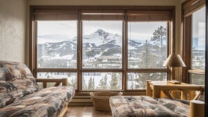 A cozy room with a large window overlooking a snowy mountain landscape. There is a sofa and a small table with a lamp.
