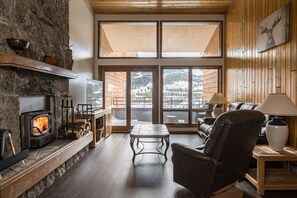 A cozy living room with a stone fireplace, a comfortable leather chair, a wooden coffee table, and large windows offering a view of the outdoors. - Mountain Condo with Pool Table and Lone Peak Views | 5 Minutes to Ski (Big Sky)