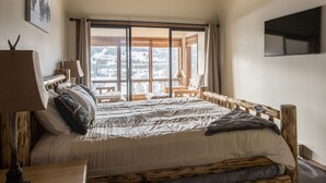 A bedroom with a large bed, a wooden headboard, a nightstand, a lamp, and a view of a snowy landscape through a large window.