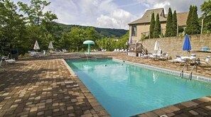 Una piscina rectangular con borde de azulejos, rodeada de sillas de descanso y sombrillas, con árboles y un edificio de fondo.