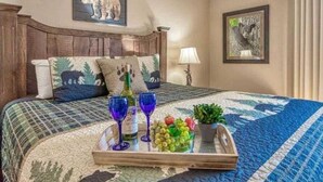 Quarto de hotel com cabeceira de madeira, cama com colcha acolchoada, duas almofadas, bandeja com copos e frutas, e quadro na parede.