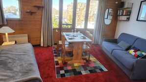 A cozy room with a dining table, chairs, a sofa, a bookshelf, and a view of buildings through large windows.