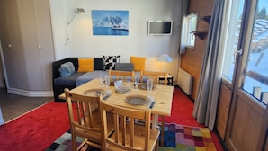 A cozy living room with a dining table set for four, a sofa with colorful cushions, a mounted TV, and a large window with curtains.