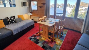 A cozy room with a sofa, a dining table with chairs, a TV, and large windows offering a view of a snowy landscape.