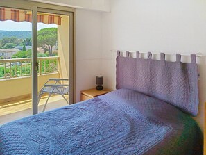 Ein Schlafzimmer mit einem Bett, einem Nachttisch, einer Lampe, einem Fenster mit Blick auf einen Balkon und einer Tür.