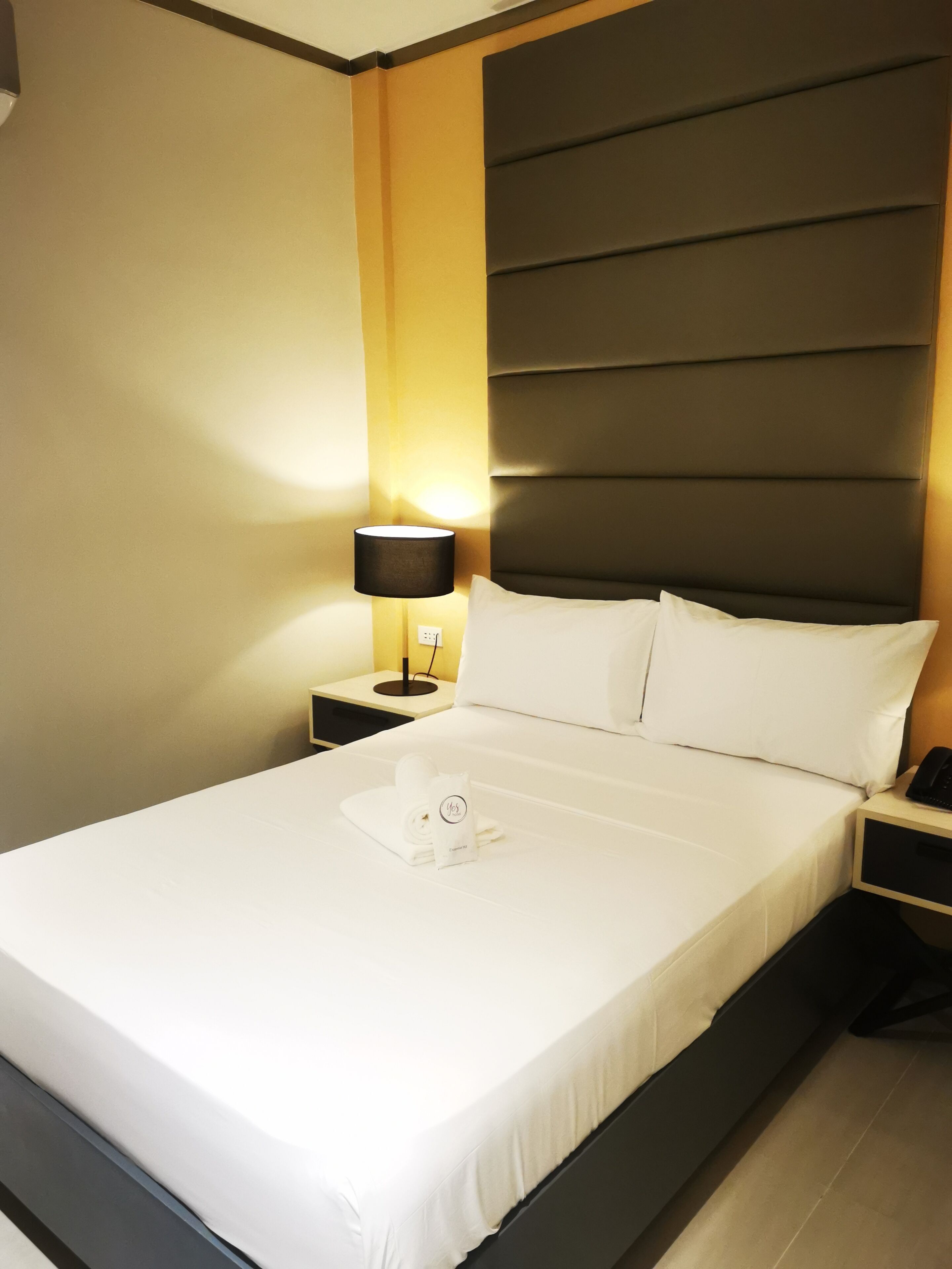 A neatly made bed with white linens, a bedside lamp, and a towel with a logo on the nightstand.