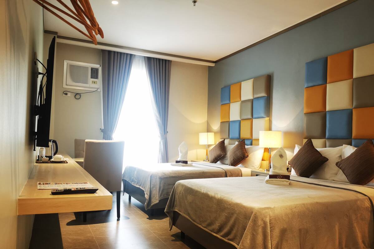 A hotel room with two beds, a desk with a chair, a coffee machine, and a window with curtains.