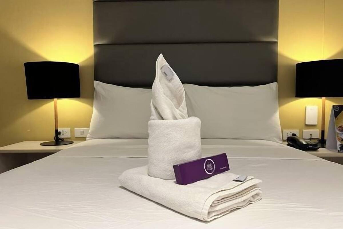 A neatly made bed with a folded towel and a purple card, two black lamps on nightstands, and a large headboard.