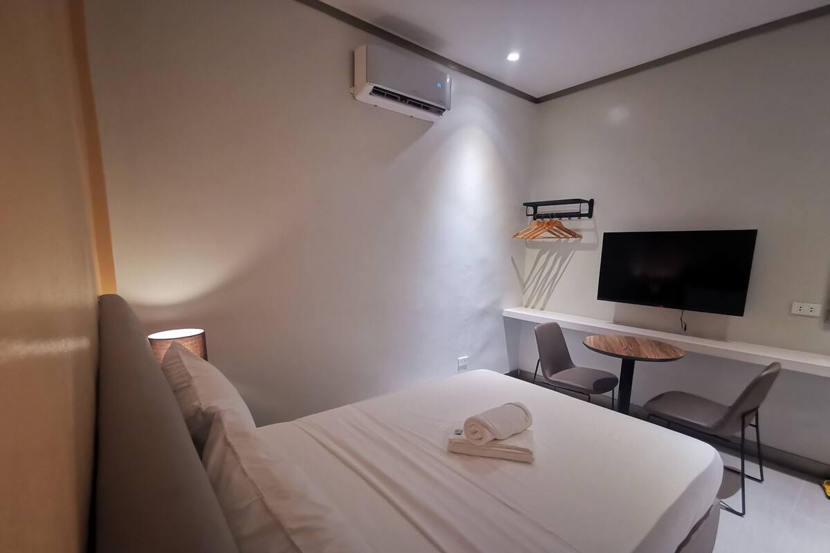 A modern hotel room with a bed, a desk, a chair, a TV, and an air conditioning unit.
